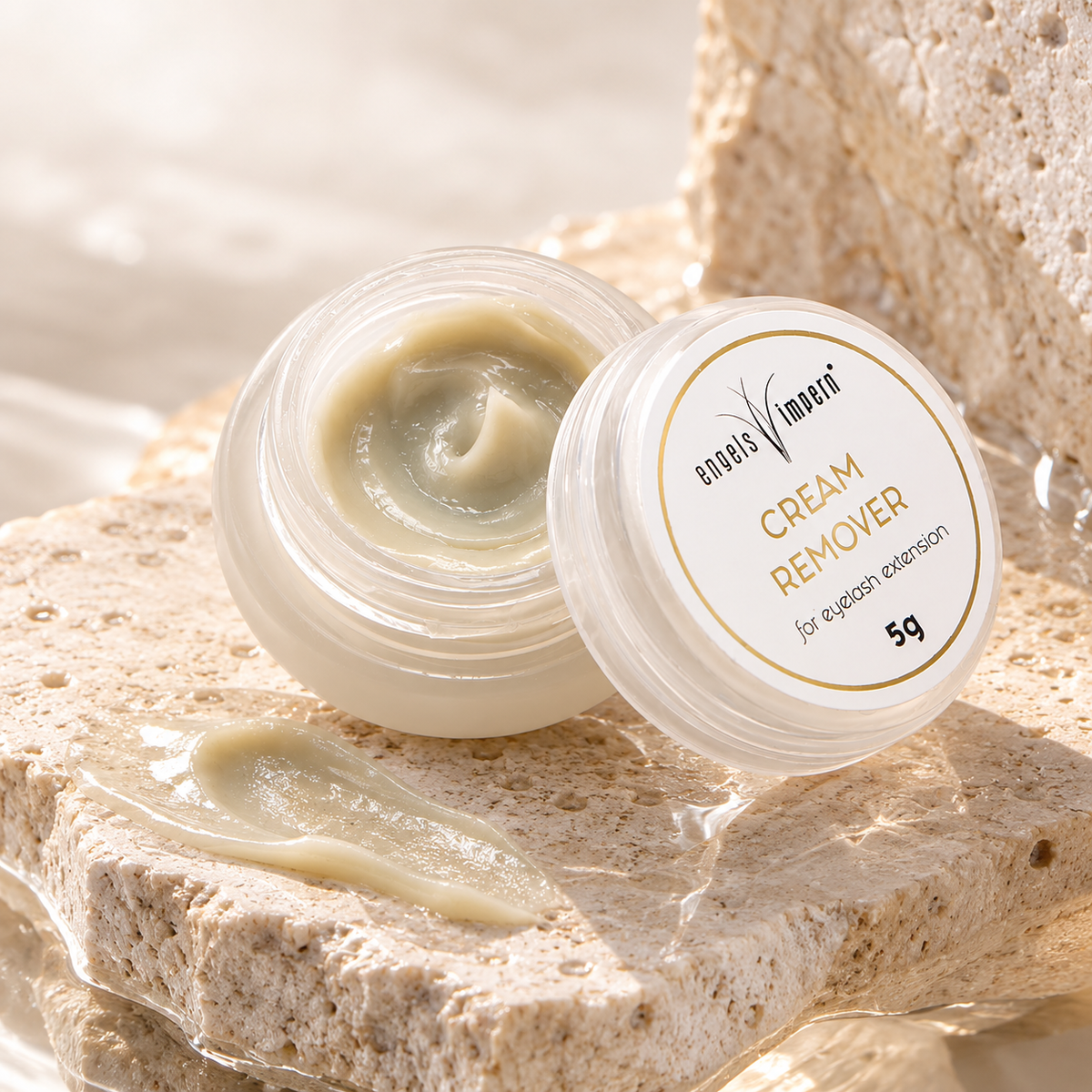 Geöffneter EngelsWimpern Cream Remover, 5 g, auf hellem Naturstein mit angelehntem Deckel und einer verstrichenen Cremeprobe im Vordergrund.