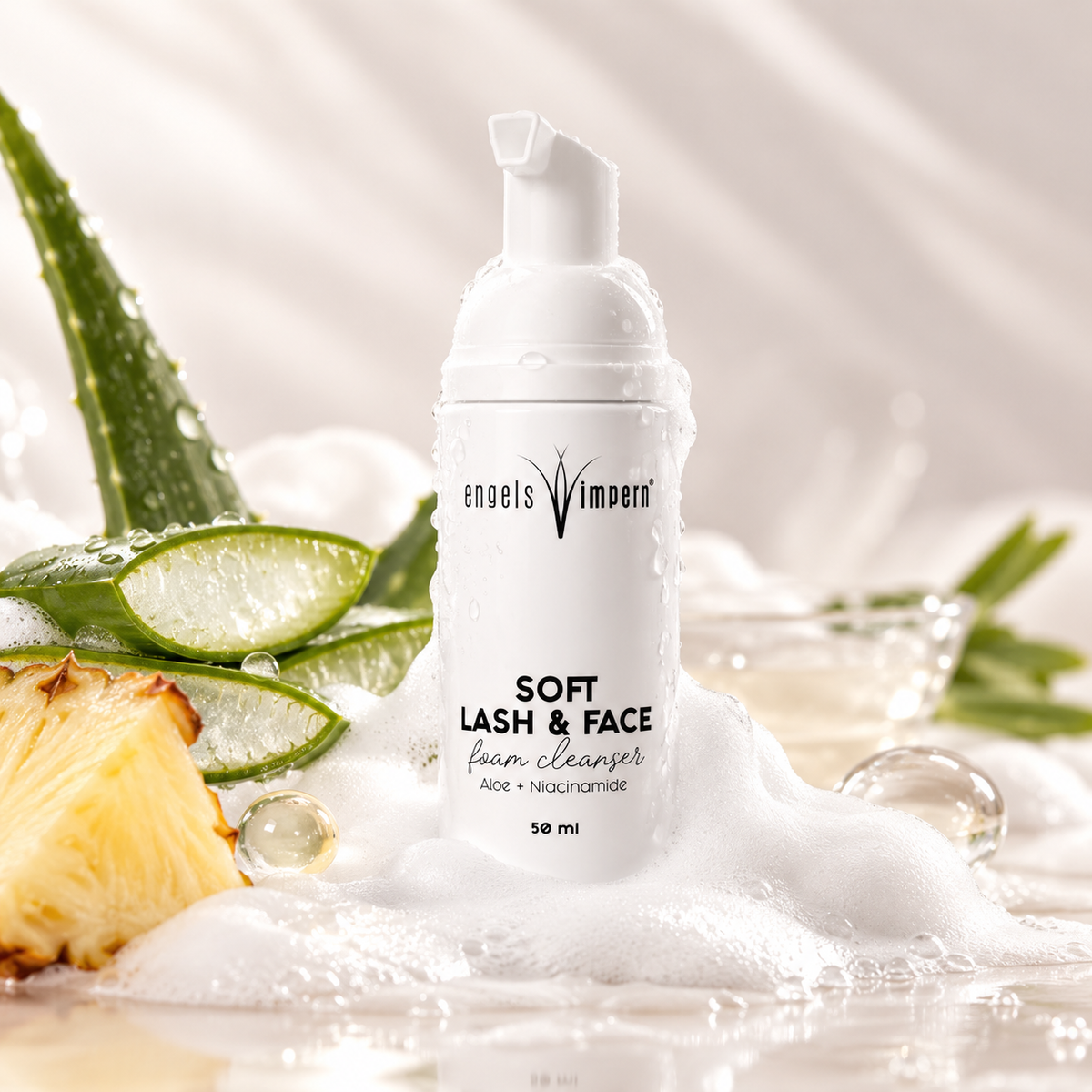 EngelsWimpern Soft Lash & Face Wimpernshampoo in einer weißen Pumpflasche, umgeben von Aloe-Blättern, aufgeschnittener Ananas, weißem Schaum und dekorativen Flüssigkeitstropfen.
